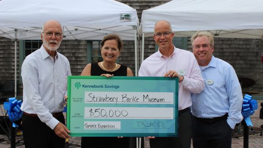 Representatives from Kennebunk Savings and Strawbery Banke hold a check for 50,000 dollars in front of a tent with instruments set up for an outdoor concert.