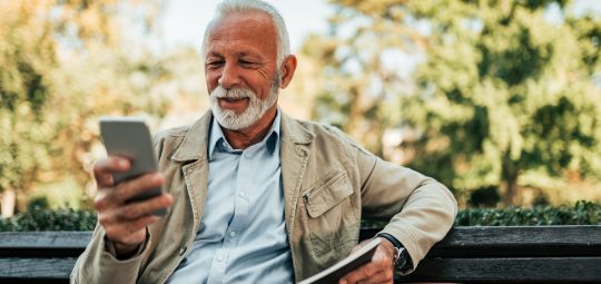 Senior man reading online news on smartphone outdoors.