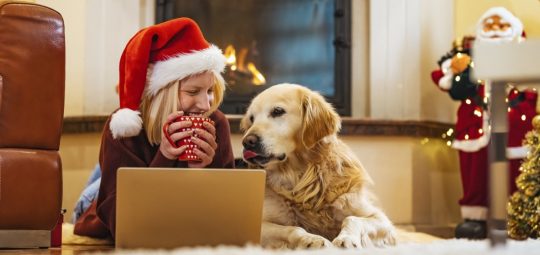Woman laying on her living room floor with her laptop and her dog. The woman is wearing a holiday hat.