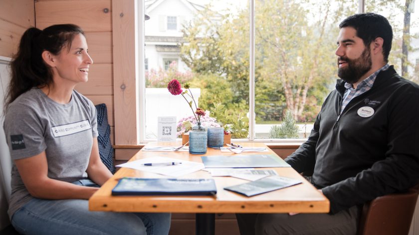 Two people sit across from each other at a wooden table in a cafe, engaged in conversation with paperwork between them.