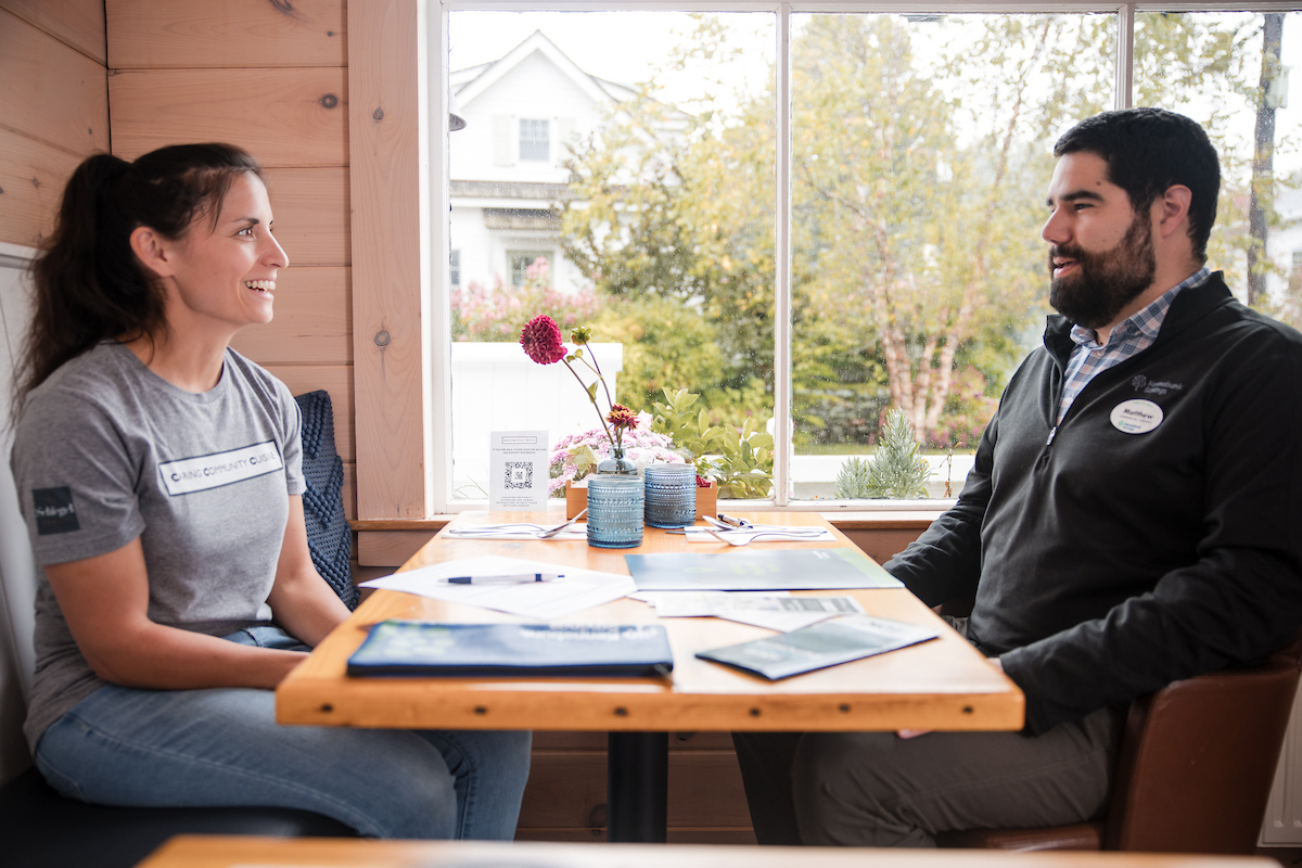 Two people sit across from each other at a wooden table in a cafe, engaged in conversation with paperwork between them.